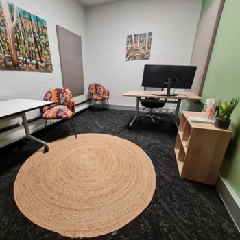 Small office with a desk, computer monitor, two patterned chairs, wall art, and a circular woven rug on a dark carpeted floor.